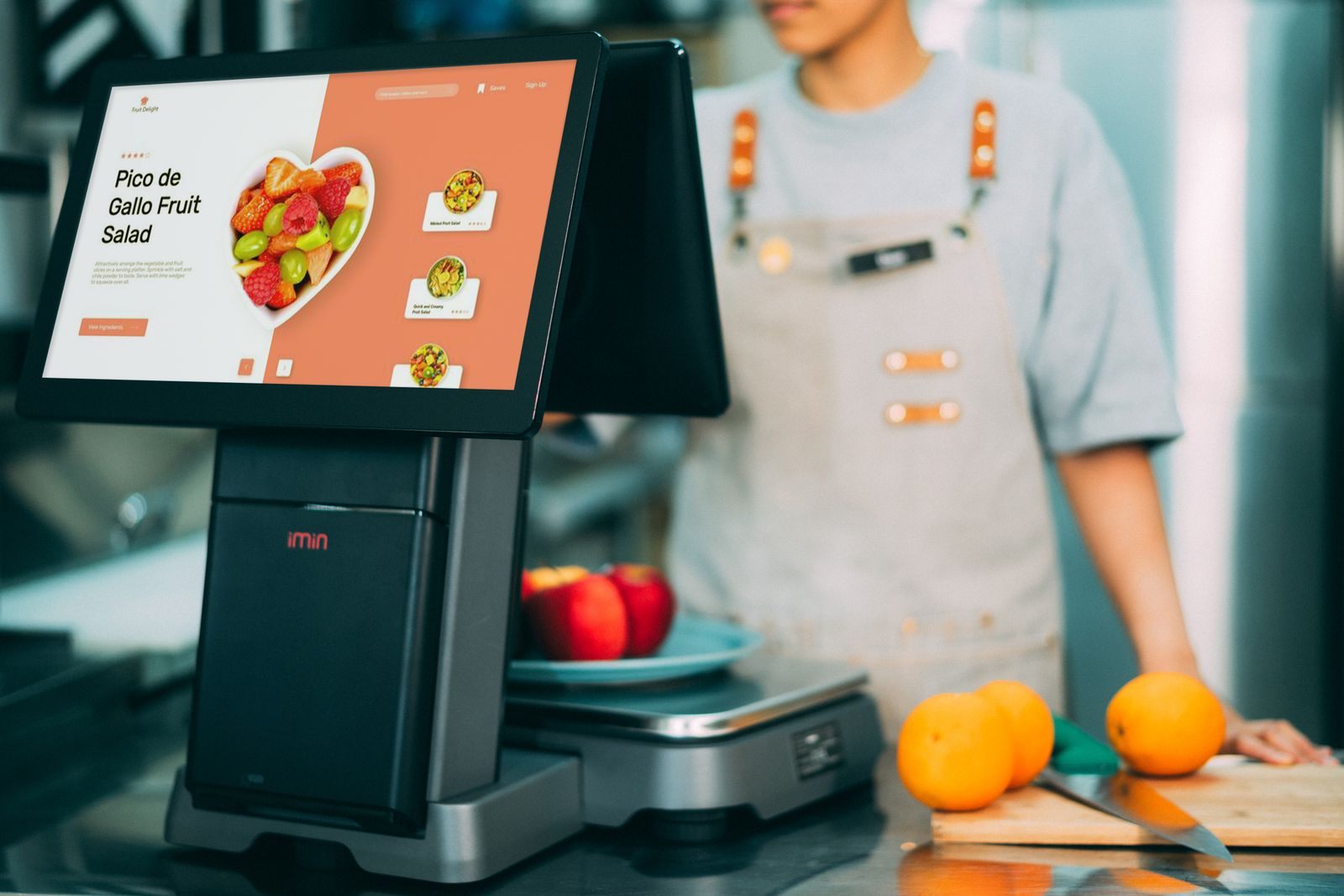 Digital checkout system in use at a grocery store with fresh fruits and a cashier in sight